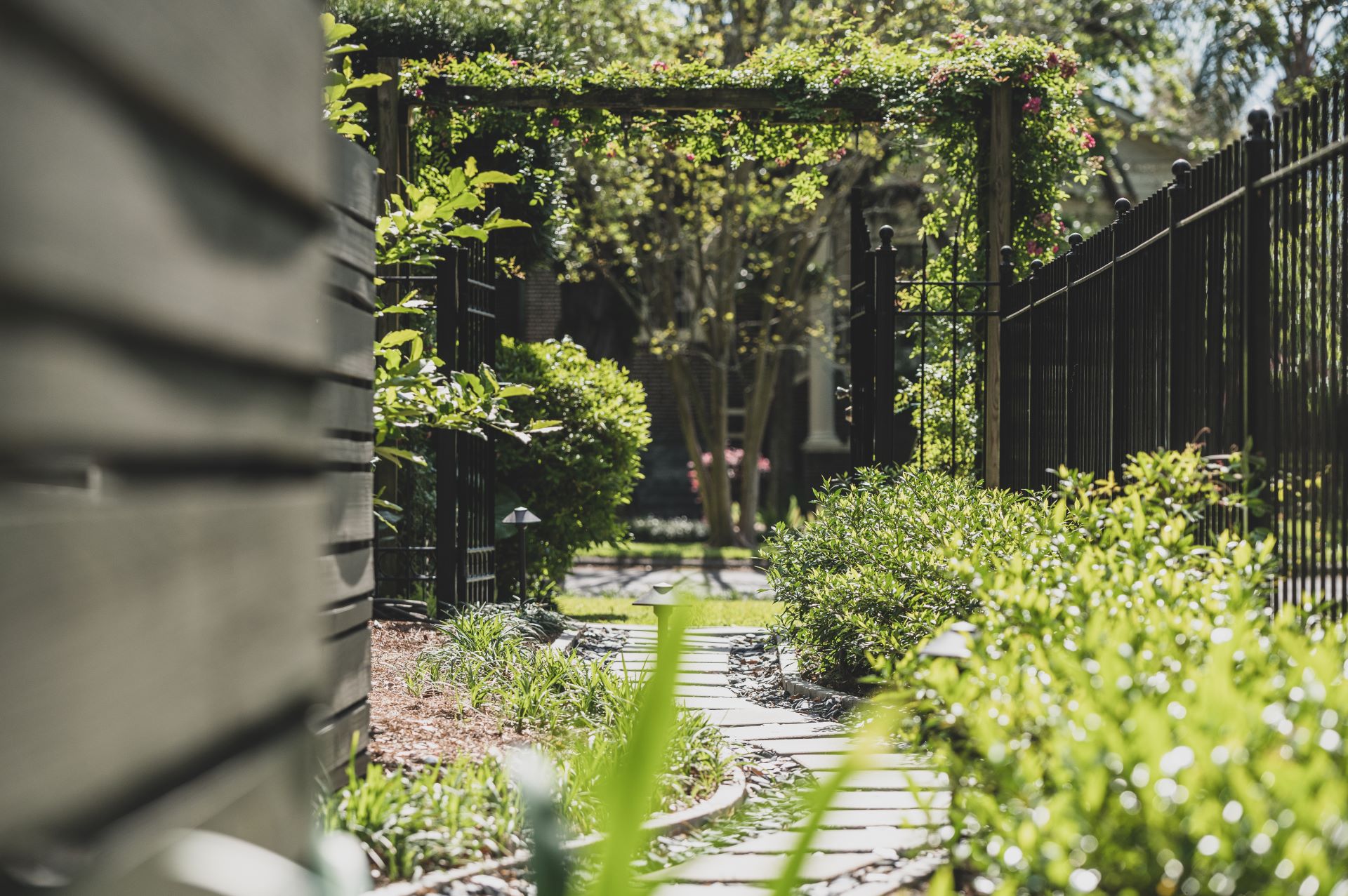 A sunlit garden pathway made of stone slabs is surrounded by lush green plants and bushes, showcasing exquisite Landscape Design New Orleans style. The path leads towards a wooden arbor draped with climbing plants. Black metal fences line both sides of the garden path, with trees and foliage in the background.