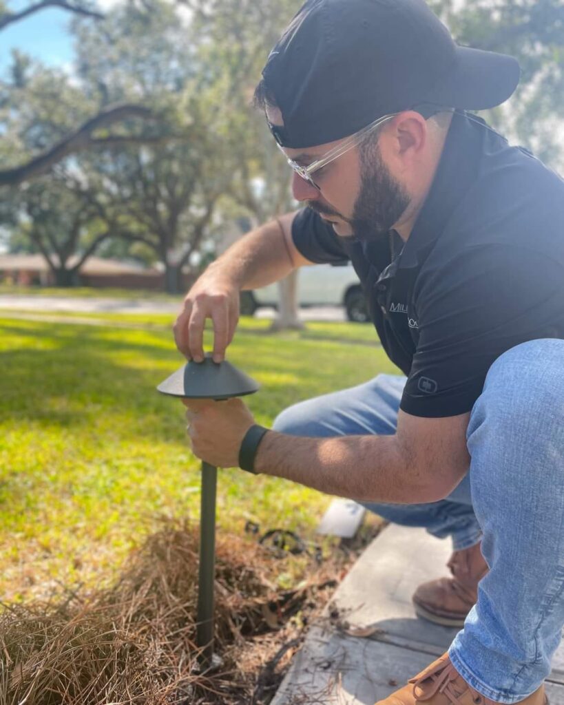 A man wearing a cap, safety glasses, and a black polo shirt is kneeling on a lawn. He is installing a small outdoor light fixture into the ground along a pathway. Sunlight filters through the trees in the background, showcasing attention to detail that defines our company values in landscape lighting solutions.