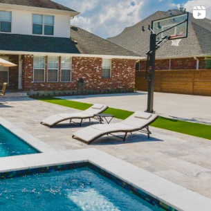 A modern backyard features a swimming pool with a spa, surrounded by a stone-tiled deck. Two reclining lounge chairs are positioned nearby. Incorporating landscape design New Orleans style, there is a basketball hoop on the deck, and in the background, there is a two-story house with a mix of brick and white siding.