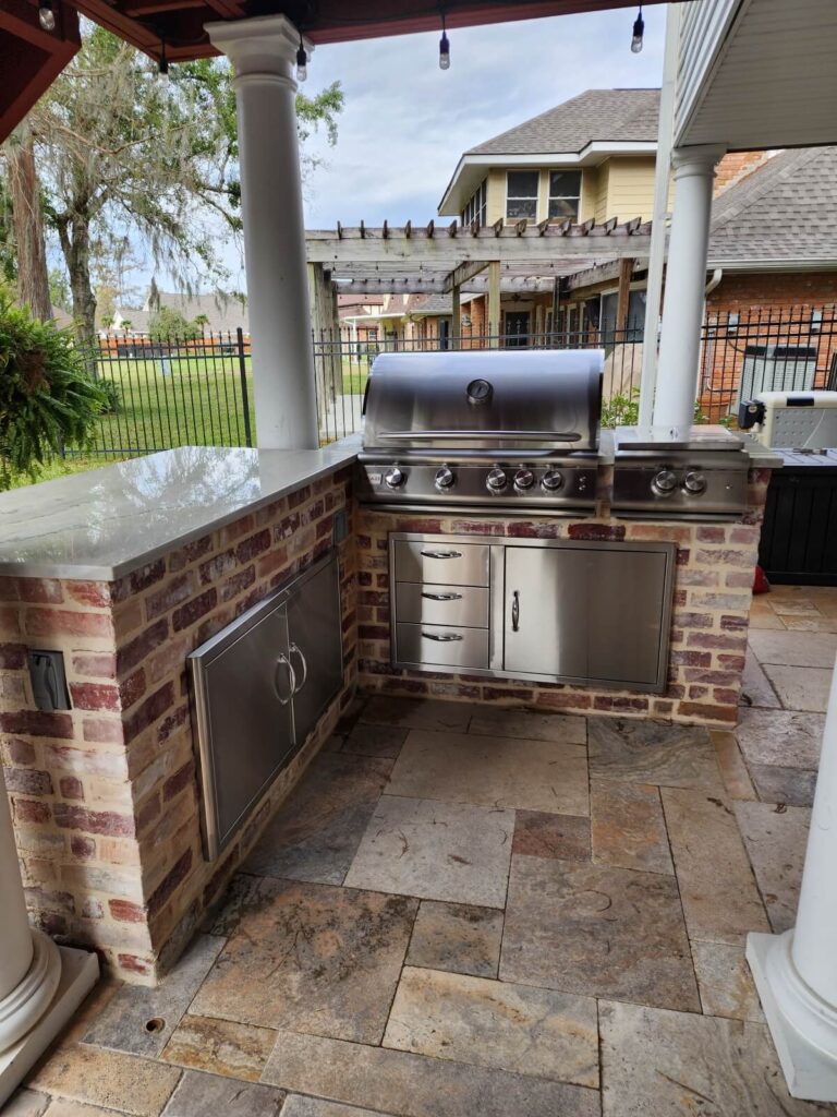 An outdoor kitchen setup featuring a stainless steel grill with multiple burners and storage cabinets built into a brick countertop. The space is surrounded by stone flooring and has a pergola in the background, offering a view of the landscaped yard and neighboring houses—a true reflection of our company's dedication to quality.