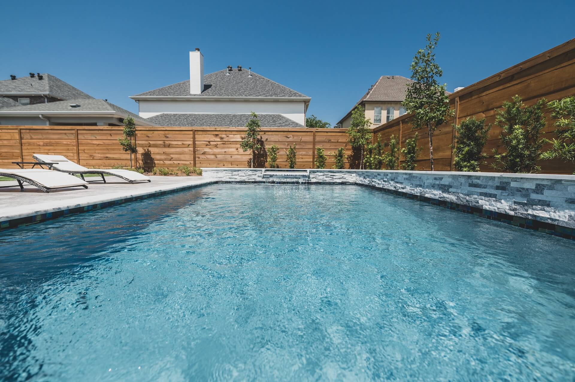 A tranquil swimming pool in a backyard with clear blue water, epitomizing refined landscape design New Orleans style. Surrounding the pool are lounge chairs and a wooden privacy fence. In the background, well-kept suburban houses boast lush green plants along the fence, under a clear blue sky.