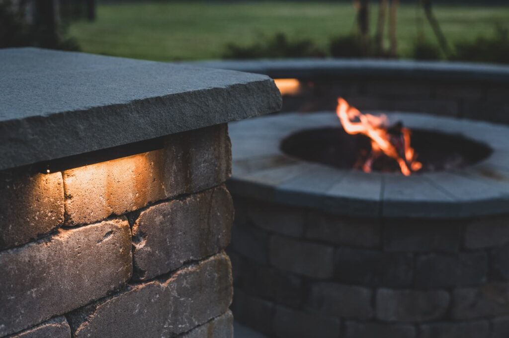 A close-up of an outdoor stone fire pit lit by ambient lighting, exemplifying exquisite Landscape Design in New Orleans. The image captures the corner of a stone structure with an embedded light on the left and a fire burning in a circular, stone-built pit in the background. The setting suggests a calm evening.