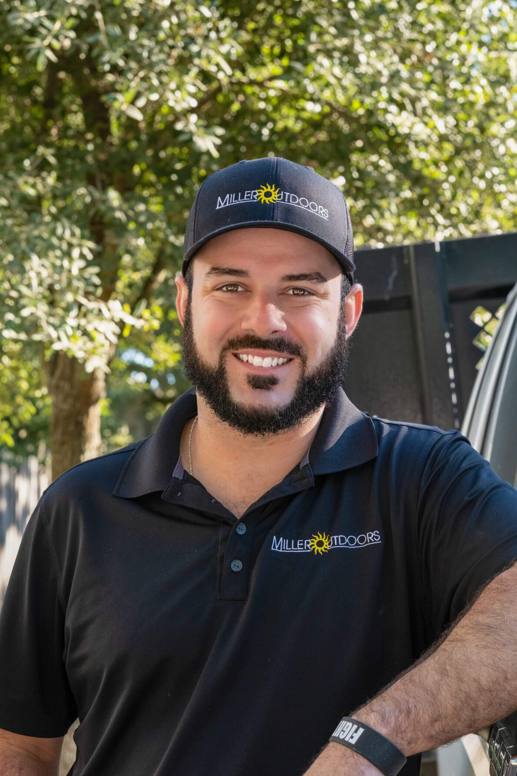 A man with a beard, wearing a black cap and black polo shirt with "Miller Outdoors" embroidered on both, stands outdoors smiling for our About Us section. Trees and a fence are visible in the background.