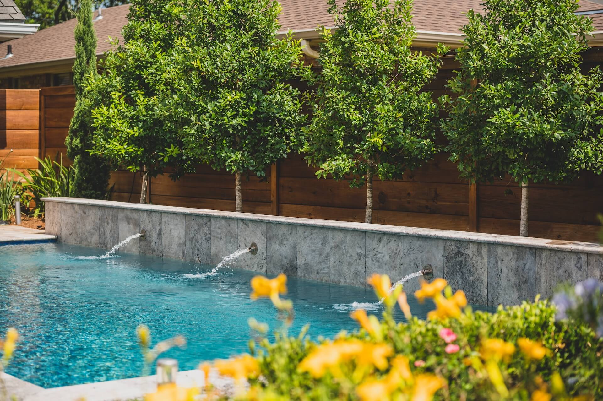 A backyard pool with a concrete wall featuring three small fountains is surrounded by lush green trees and a wooden fence. Bright yellow flowers are in the foreground, enhancing the serene outdoor setting, showcasing impeccable landscape design reminiscent of New Orleans charm.