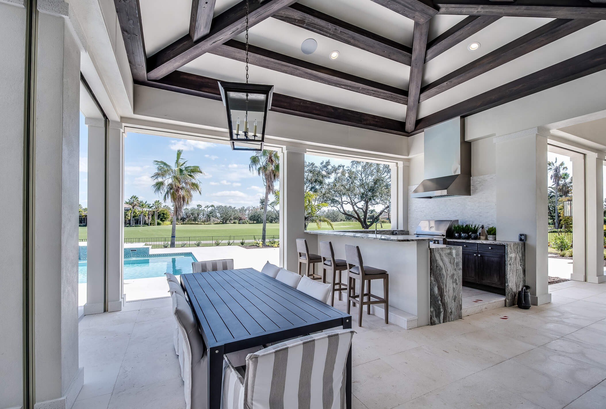 An outdoor patio featuring a long dining table with striped cushioned chairs, a bar area with barstools, and a modern kitchen setup. The ceiling has exposed wooden beams—exemplifying the landscape design process—and the patio opens to a view of a swimming pool, lush greenery, and palm trees.