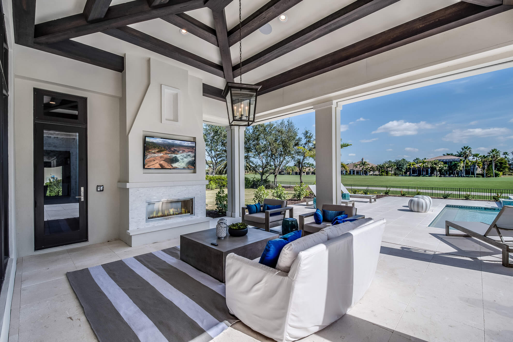 A stylish outdoor patio with a white couch, blue cushions, a coffee table, and a striped rug faces a large fireplace with a TV mounted above. The space, inspired by Landscape Design New Orleans, opens to a pool area and a manicured lawn with trees and a clear blue sky in the background.