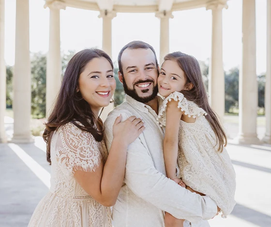 A family of three poses for a photo in an outdoor pavilion with columns. The mother and father smile at the camera while the father holds a young girl who smiles sweetly. They are all dressed in coordinated light-colored clothing, embodying the essence of our "About Us" section.