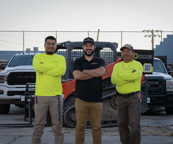 Three men stand confidently in front of construction vehicles. Two men wear bright yellow long-sleeve shirts, and the man in the middle wears a black shirt and hat. They are outside in a parking lot or work area with industrial trucks and equipment, showcasing promising job opportunities in their careers.