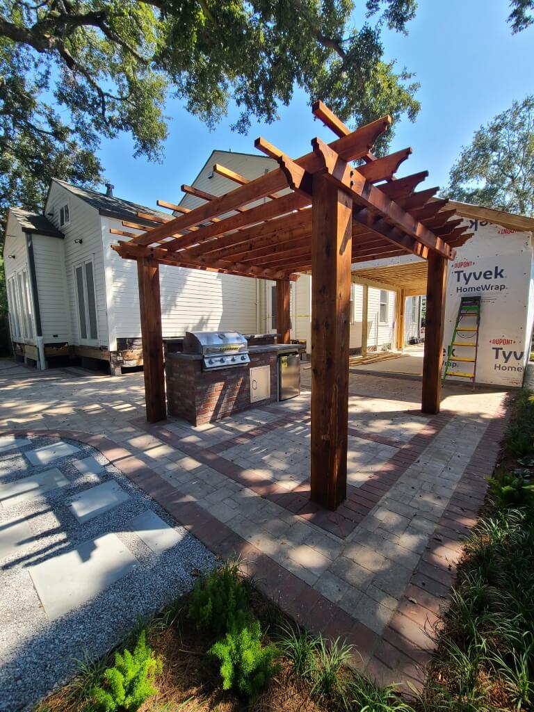 A partially constructed outdoor kitchen at the residence features a brick and metal grill setup under a wooden pergola. The ground is paved with a mix of bricks and gravel pathways. The scene is set against two white buildings, with one wrapped in Tyvek HomeWrap, under a sunny sky.