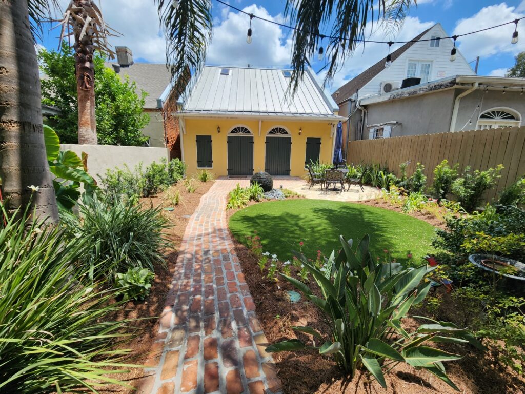 The Aceto Residence features a garden with a red-brick pathway leading to a yellow building with a silver roof and three green doors. The lush garden boasts various plants and flowers, with string lights hanging overhead. Two outdoor chairs and a table are placed along the path, inviting relaxation.