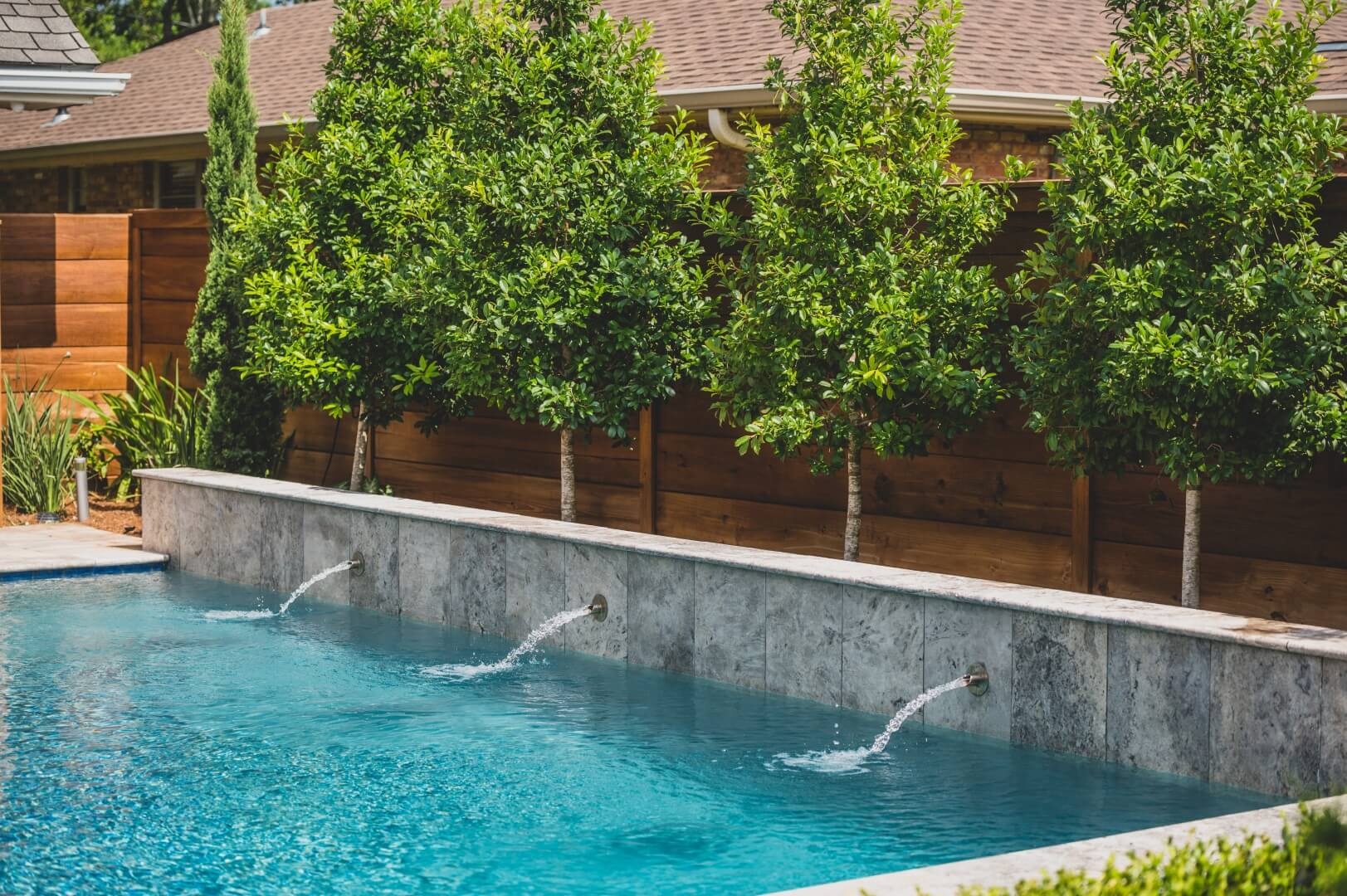 The Naylon Residence boasts a backyard pool with clear blue water and three water spouts flowing into it. It is bordered by a light gray stone wall and lush green trees, with a wooden fence in the background. The scene is well-lit with natural sunlight, creating a perfect oasis.