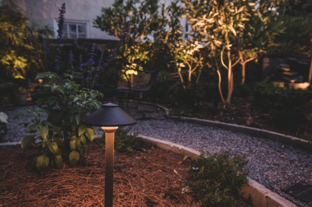 A garden pathway illuminated by a small black outdoor lamp. The path is lined with plants and trees, creating a serene and softly lit atmosphere in the evening. The ground is covered with mulch and the pathway with gravel, dispelling common landscape design myths about complexity.