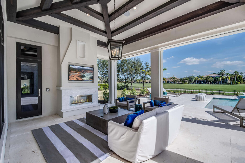 A luxurious outdoor patio features a comfy white sofa with blue pillows and a large striped rug. The area is equipped with a fireplace, mounted TV, and dining table. Beyond the patio, debunking common landscape design myths, there’s a pool, golf course, and scenic view of trees under a clear blue sky.