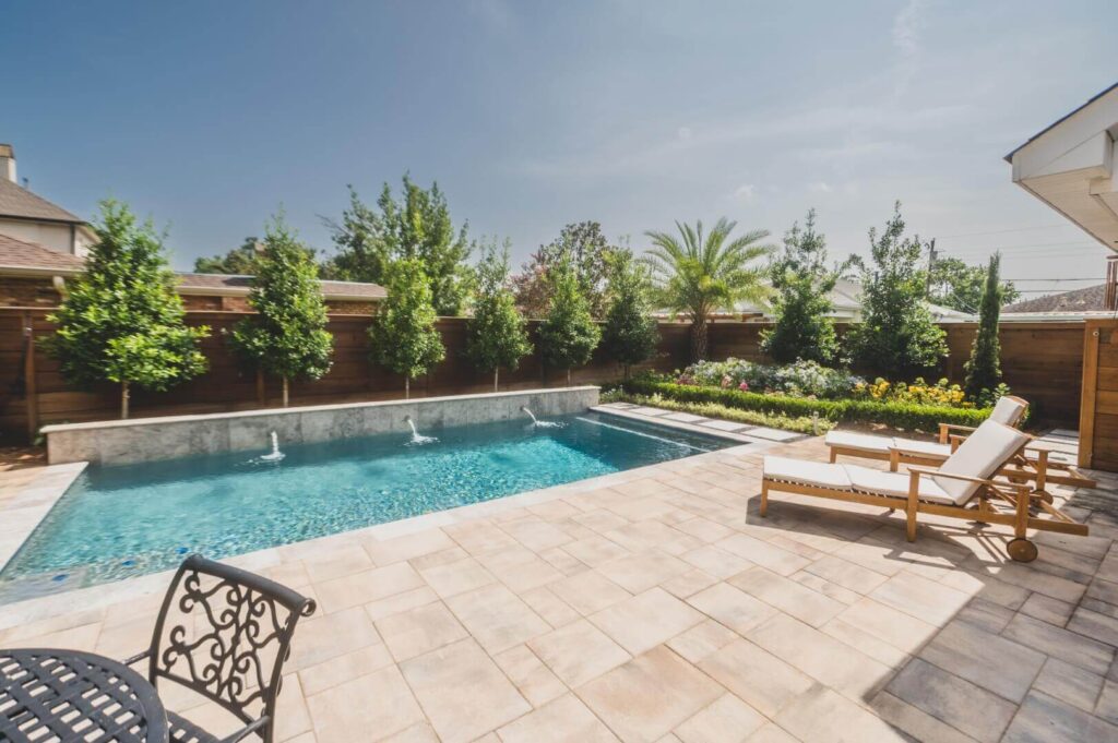 A backyard swimming pool surrounded by a tiled patio area reveals a space debunking landscape design myths. Lush trees and shrubs line the wooden fence in the background. Two wooden lounge chairs with white cushions sit by the pool, with a round metal table and chairs on the left side.