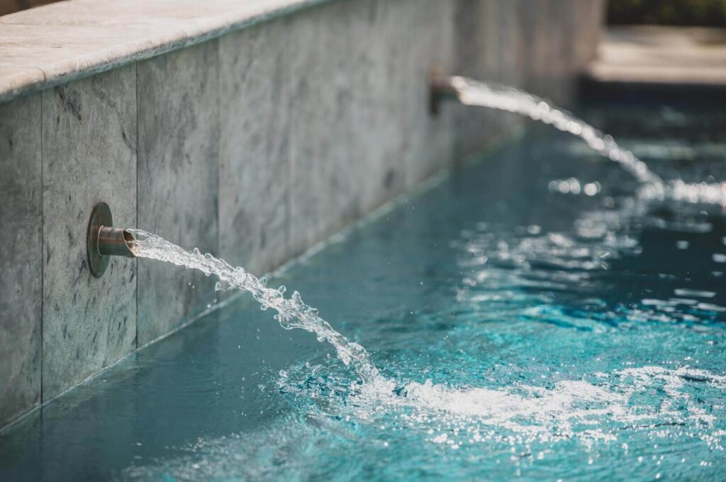 Water flows from two spouts set into a stone wall, spilling into a serene, clear blue pool. The surface of the pool is gently rippling, and the thoughtfully crafted landscape design dispels any myths about creating truly tranquil outdoor spaces. The scene conveys a deep sense of calm and relaxation.