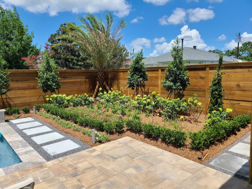 A landscaped backyard features a wooden fence, a variety of green plants, and neatly arranged flowers. A stone pathway leads through the garden, and part of a swimming pool is visible in the foreground. With such beauty on display, one might wonder: How much does landscape design cost? The sky is blue with a few scattered clouds.