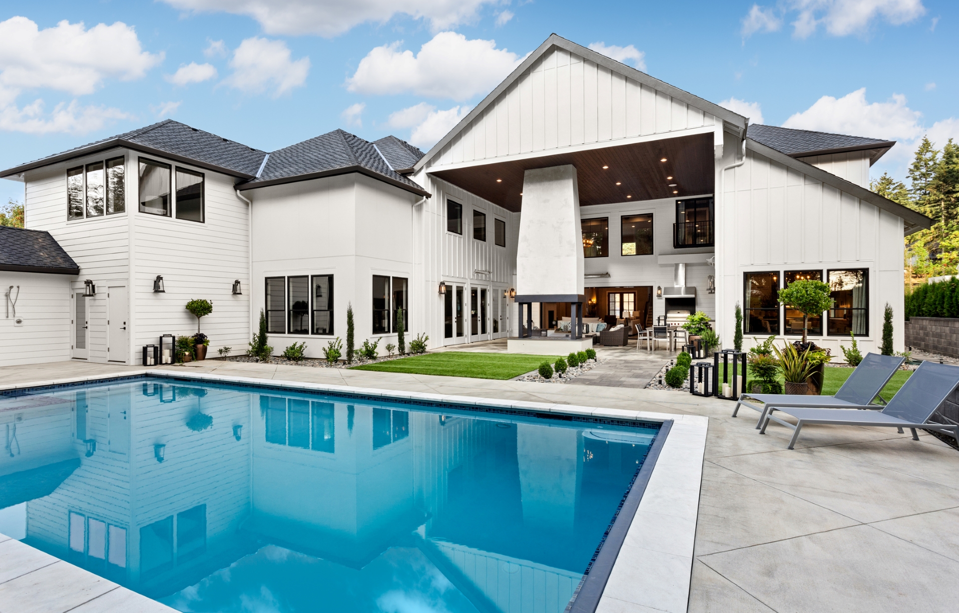 A modern, large white two-story house in New Orleans features expansive windows and a well-appointed outdoor living space. The backyard includes a rectangular swimming pool surrounded by a concrete deck, lounge chairs, and a covered patio with dining furniture. The sky is clear with a few clouds.