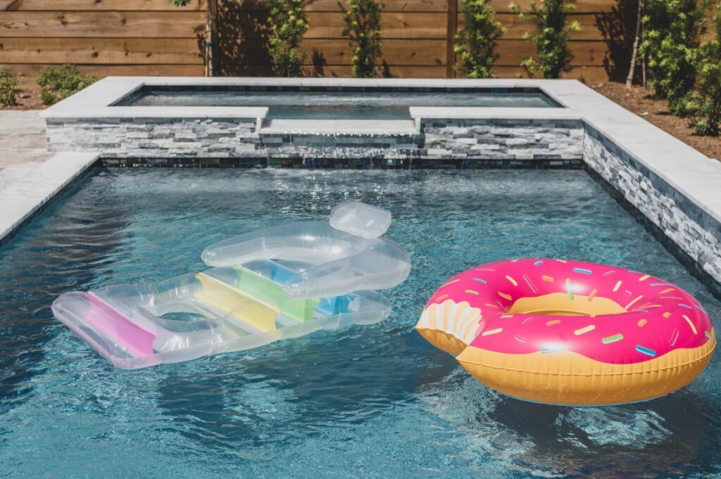 A swimming pool with two inflatable floats, one resembling a pink donut with sprinkles and the other a clear lounger with colorful stripes. The pool, inspired by Pool Design New Orleans, has a waterfall feature and is surrounded by stone walls and lush greenery.