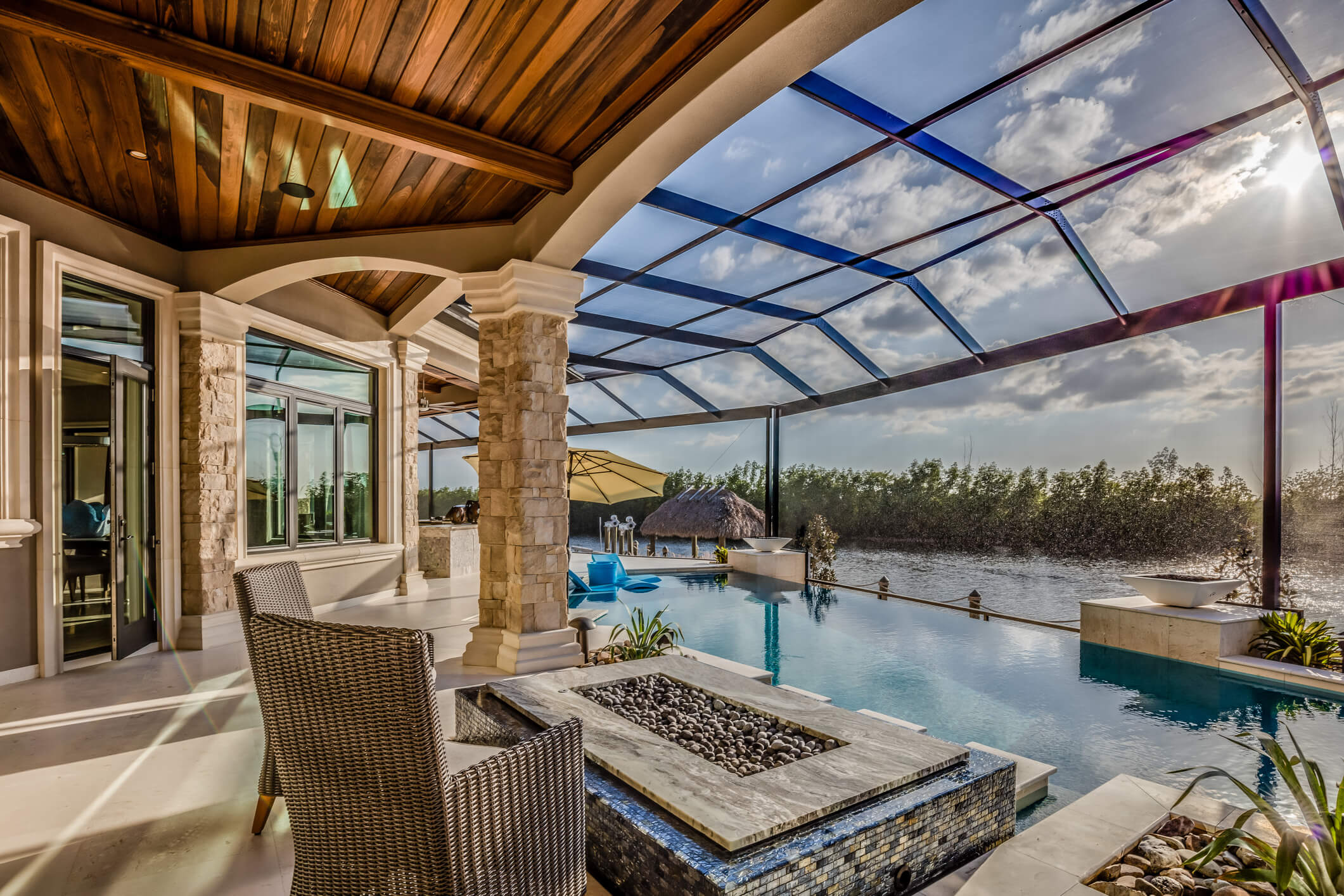 A luxurious poolside area with wicker chairs and a fire pit offers a Pool Design New Orleans charm. The infinity pool extends into a scenic view of the lake, surrounded by a screen enclosure. Stone columns, a wooden ceiling, and tropical decor with a cabana and lush greenery complete this serene retreat.