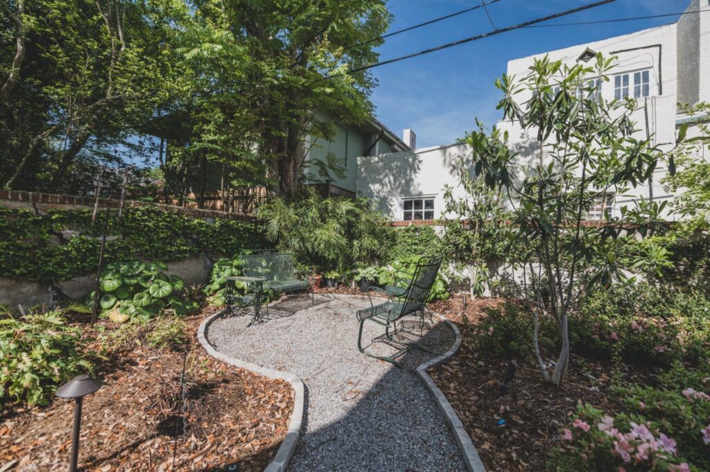 A peaceful garden with a gravel pathway leading to a seating area with two green metal chairs and a small table. The lush greenery, tall ivy-covered wall for privacy, and a white building in the background under a clear blue sky make one wonder: How much does landscape design cost?