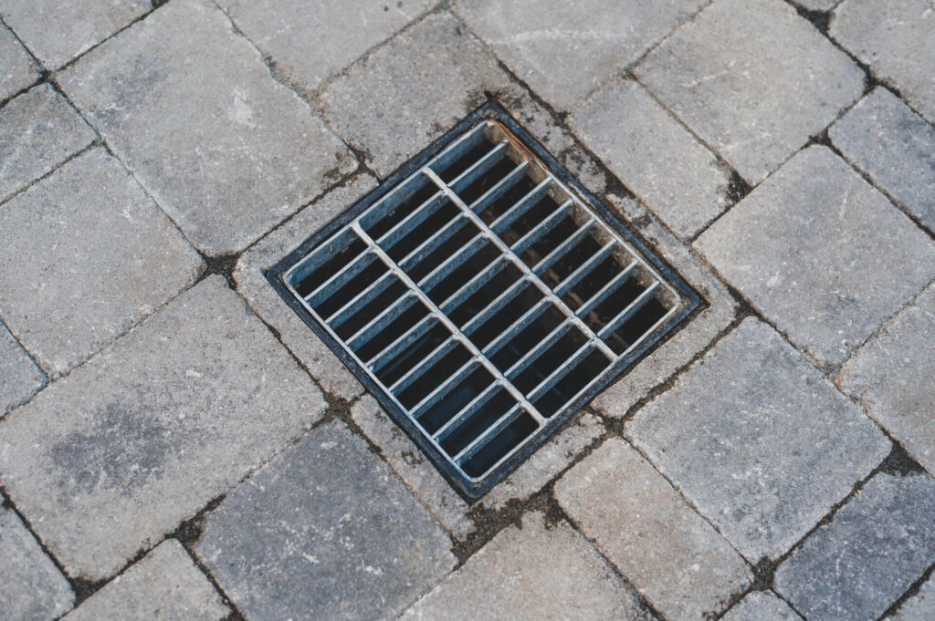 A square metal grate embedded in a cobblestone pavement at the Versailles residence. The grate covers a small drain. The cobblestones vary slightly in color, ranging from light to dark gray, and have visible dirt and wear in the grooves between them.