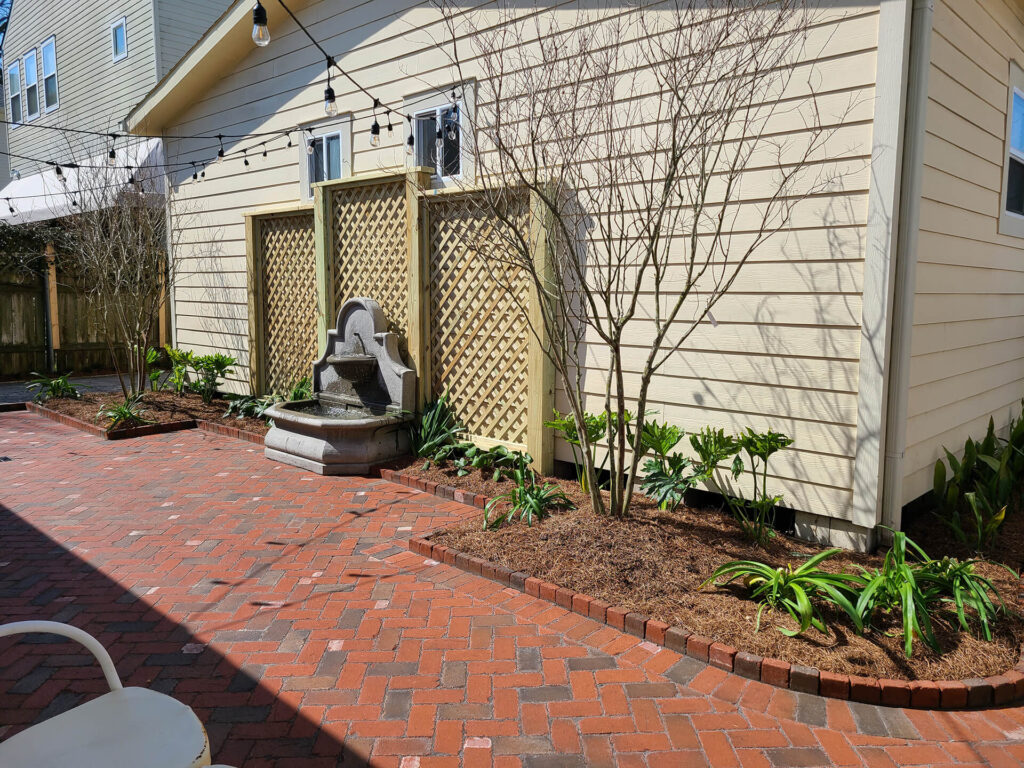 A sunny backyard at the Duxstad residence features a brick patio and raised flower beds. A tiered stone fountain is placed against a cream-colored shed with lattice panels. Overhead, string lights dangle while various green plants thrive in the beds, creating an inviting and picturesque space.