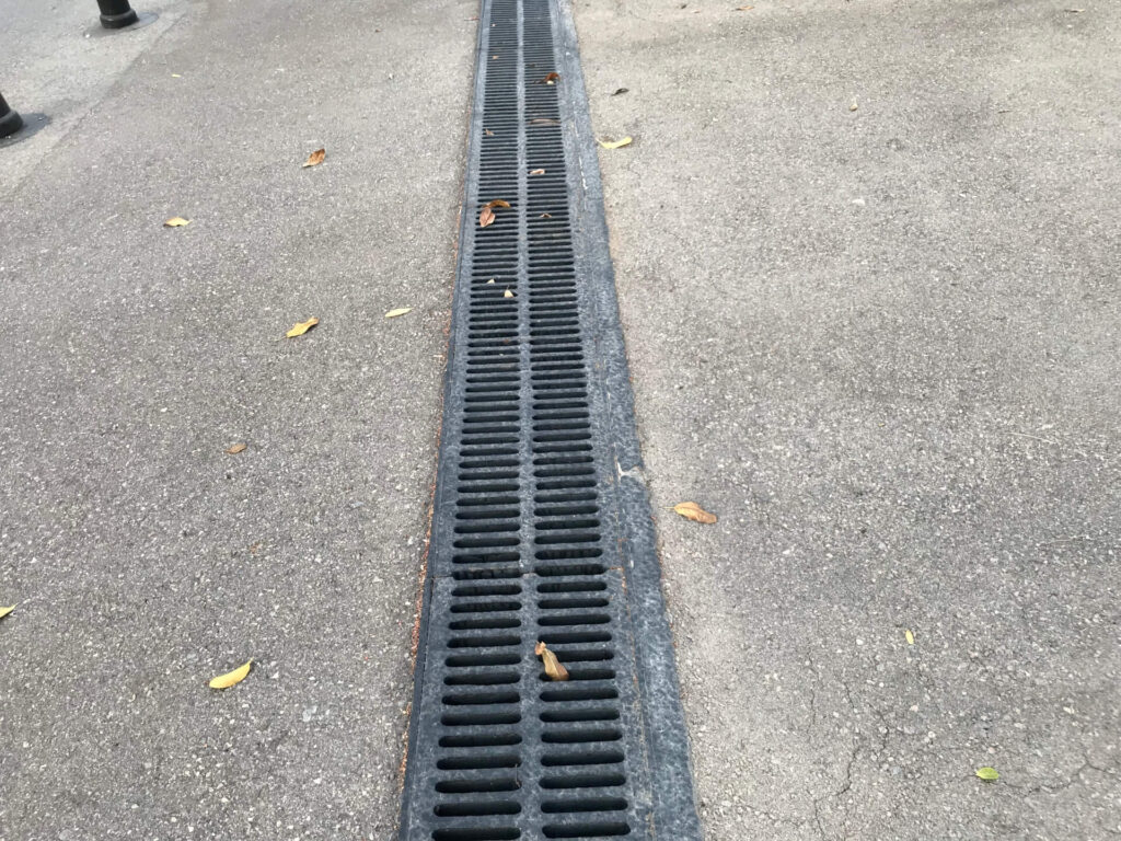 A long, black metal drainage grate, expertly installed by drainage contractors from New Orleans, runs through the middle of a paved pathway. A few scattered leaves lie on the ground with a sidewalk curb visible on the left side.