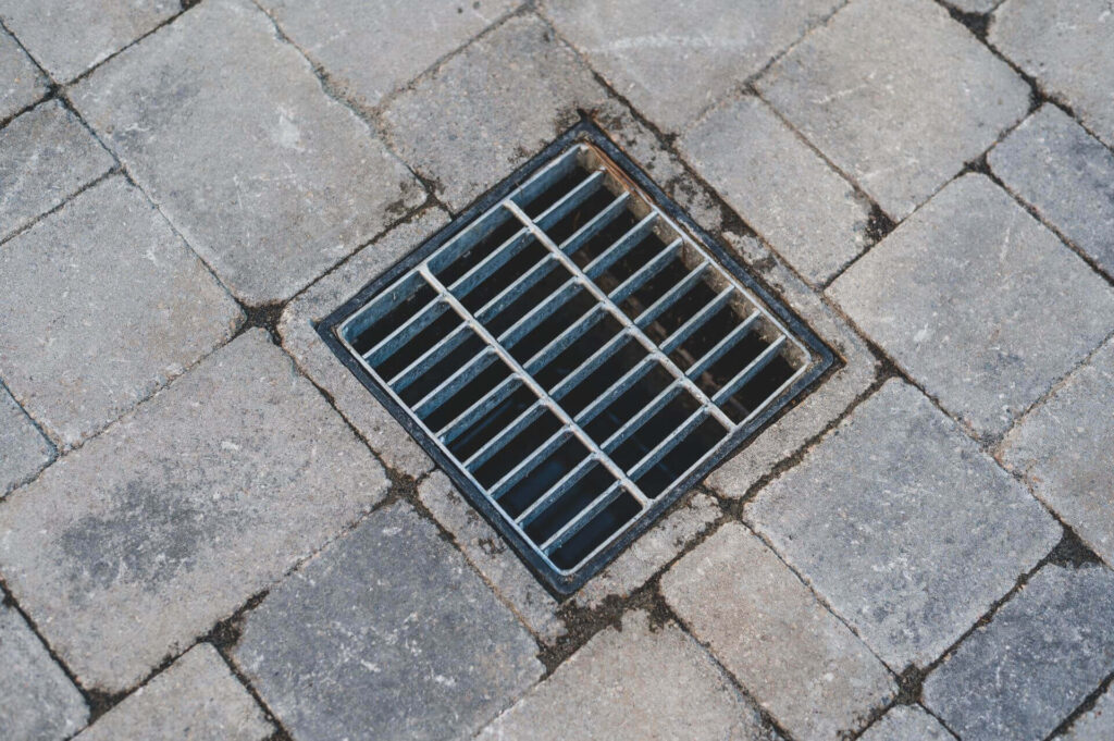 A square metal grate over a drain, likely crafted by drainage contractors in New Orleans, is embedded in a cobblestone pavement. The stones are arranged uniformly, with gaps visible between them. Evenly spaced grate bars reveal the dark interior of the drain beneath.