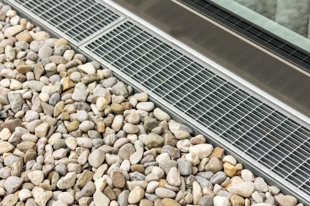 A metal grate, installed by leading drainage contractors in New Orleans, is set into the ground beside a modern building window, surrounded by evenly spread small gray and tan pebbles.