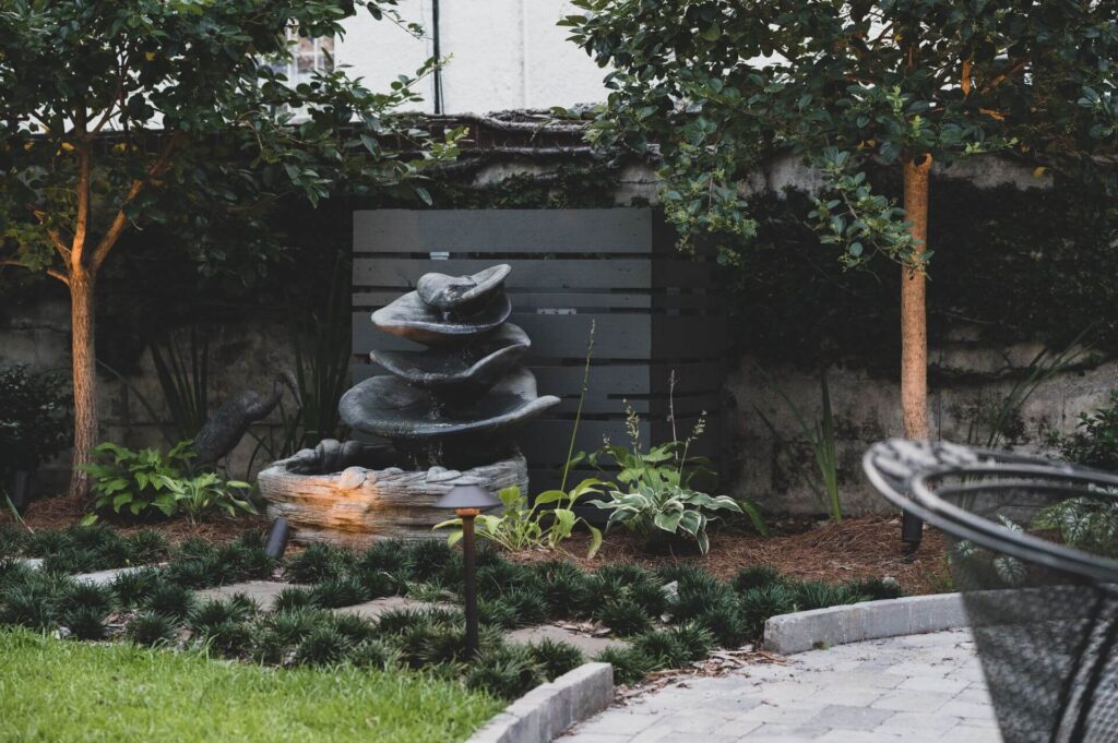 A tranquil garden features a modern stone sculpture with layered rocks. Surrounded by neatly trimmed plants and bushes, the scene is softly lit by strategically placed landscape lights near a paved area and a wire-frame patio chair in the foreground.