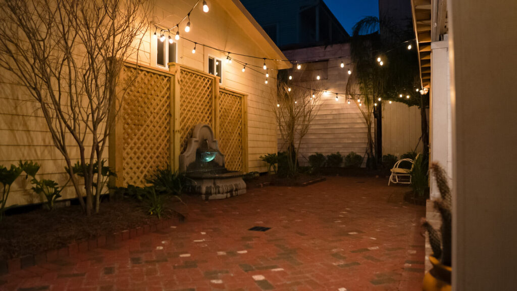 A cozy backyard patio at night with string and landscape lights illuminating the space. There is a brick floor, walls with lattice accents, sparse trees, and a stone water fountain. A white chair is visible in the dim corner, enhancing the serene ambiance.