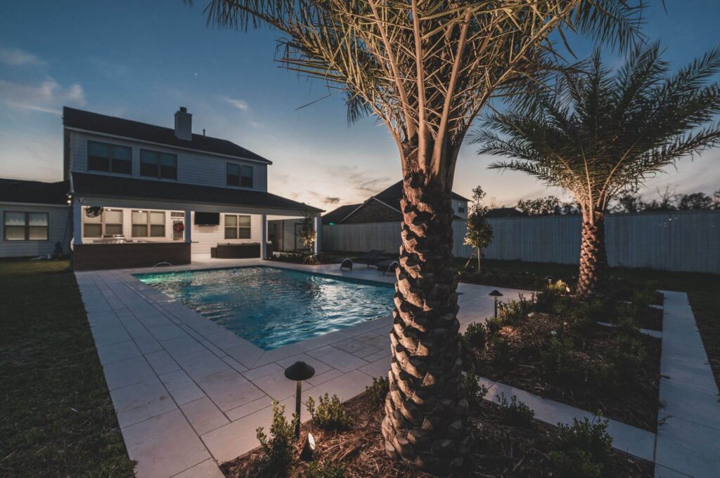 A backyard view at dusk showcases a swimming pool encircled by palm trees and pristine landscaping. Landscape lights gently highlight the modern two-story house in the background, partially illuminated to enhance its contemporary charm.