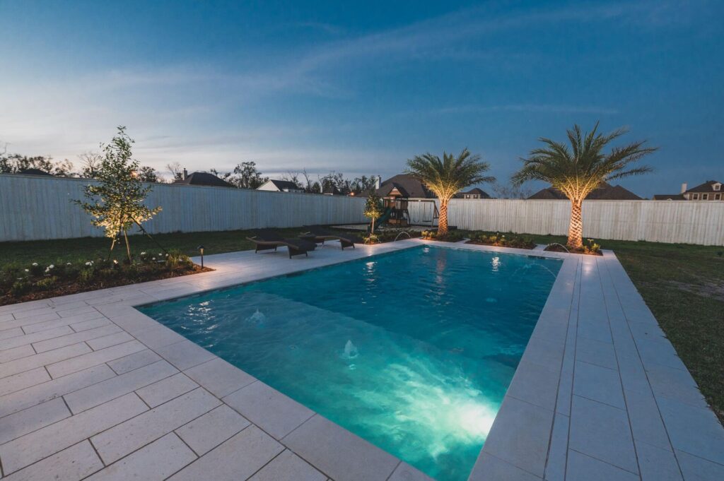 A backyard scene at dusk features a well-lit swimming pool surrounded by a stone patio. Landscape lights accentuate two lounge chairs sitting poolside, flanked by palm trees. A white privacy fence and houses are visible in the background under a darkening blue sky.