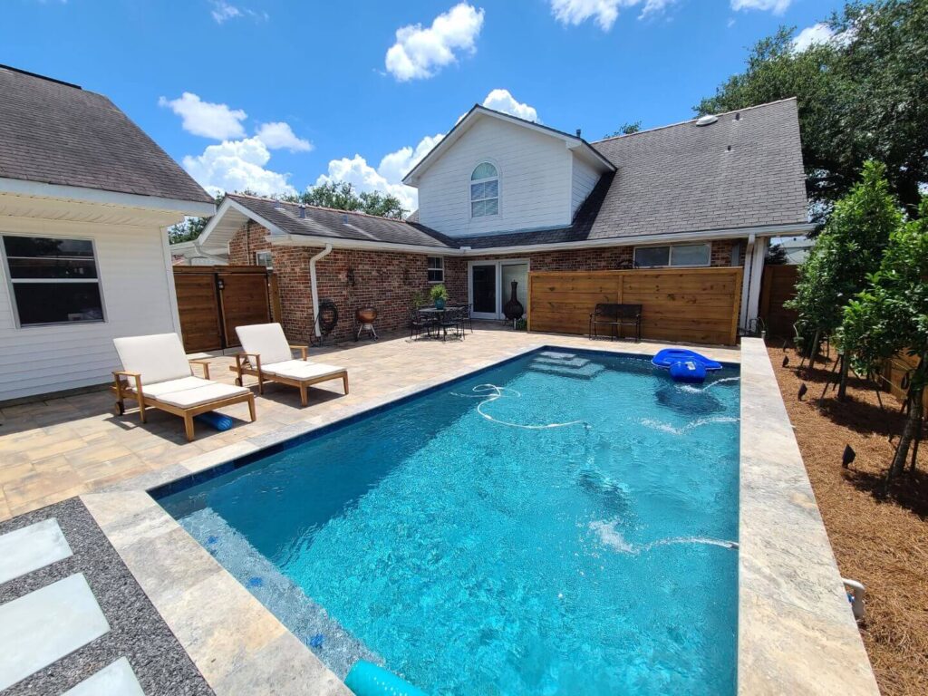 A custom saltwater pool graces the backyard, complete with a stone patio and two wooden lounge chairs adorned with cushions. A blue inflatable raft floats peacefully in the water. The house, featuring brick and white siding with a sloped roof, sits under a clear sky dotted with a few clouds.