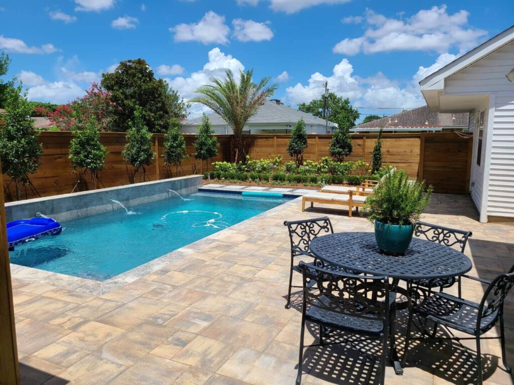 Benefits of a Custom Saltwater Pool. A backyard showcases a custom saltwater pool encircled by a patio with stone tiles. There's a black metal table and chairs, a planter, and a wooden fence with lush greenery and a palm tree. A white house is partially visible, accentuating the backyard benefits of this serene retreat.