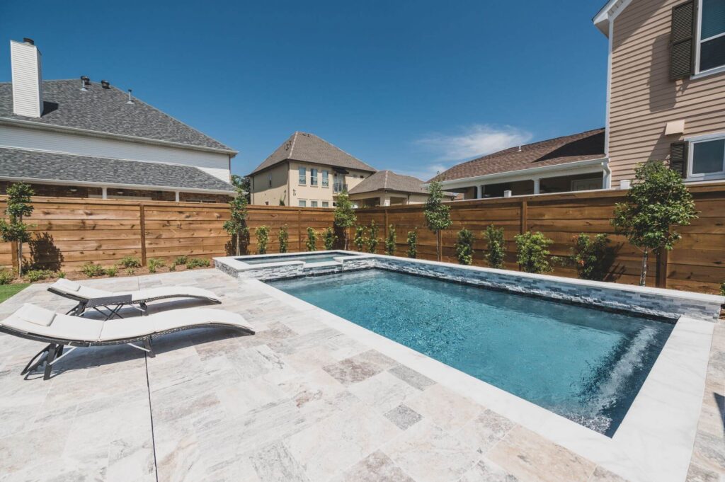 A backyard with a rectangular custom saltwater pool, surrounded by a stone patio. Two lounge chairs are on the left side. Small trees line the wooden privacy fence. Several houses are visible in the background under a clear blue sky, offering all the benefits of tranquil seclusion and convenience.