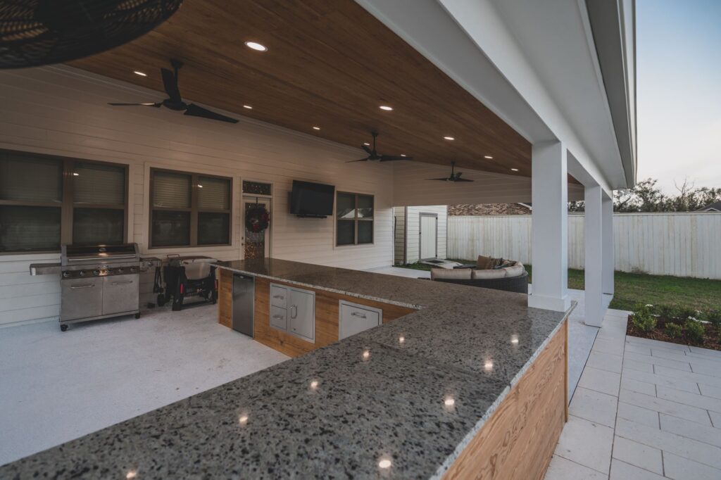 This New Orleans outdoor kitchen features a polished stone countertop bar, built-in grill, and wooden ceiling. Ceiling fans and recessed lights hover above, casting a glow on the scene. Enjoy the wall-mounted TV from the couch, with a fenced lawn visible in the background.