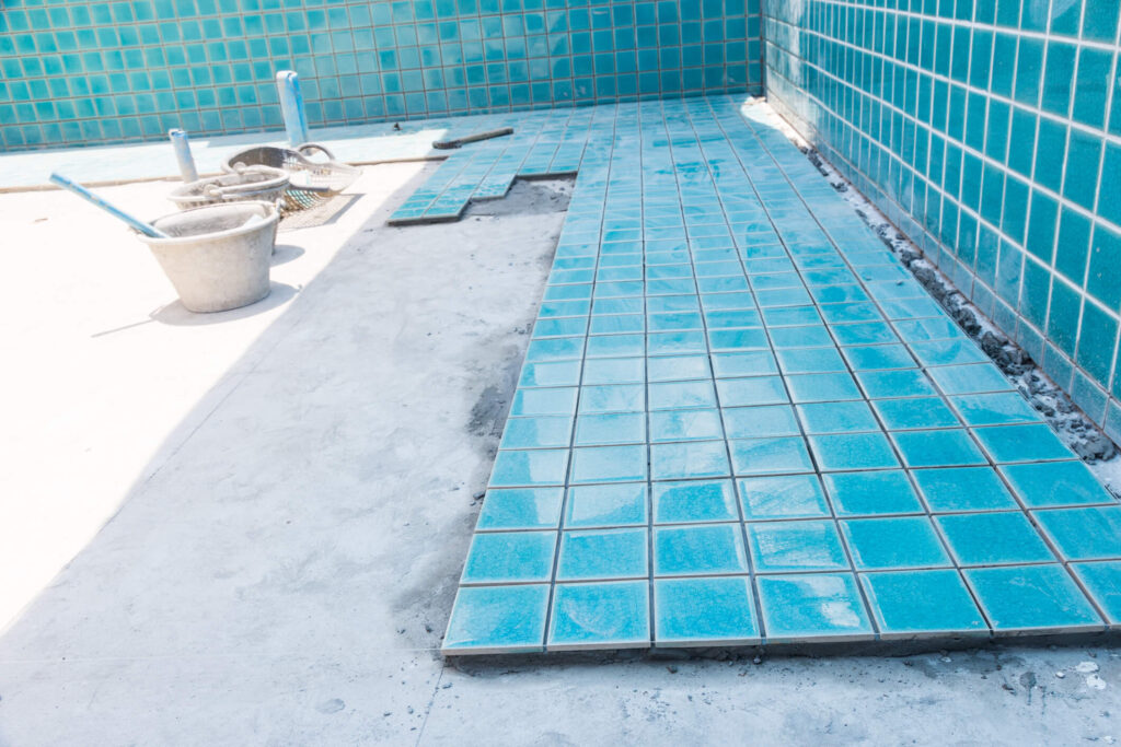 A swimming pool under construction showcases a section of bright blue tiles being artfully laid on the floor, reflecting the careful choice of the right pool tile. Nearby, a bucket and construction tools rest on the unfinished concrete surface, while matching blue tile lines adorn the pool walls.