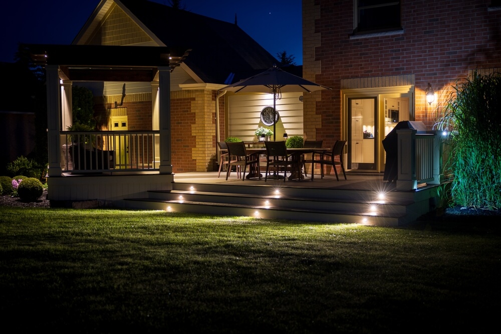 A backyard patio at night with soft New Orleans landscape lighting. There is a wooden deck with steps leading to a table set under an umbrella. A pergola with more seating is on the left. The scene is warmly lit, highlighting the brick and siding of the house.