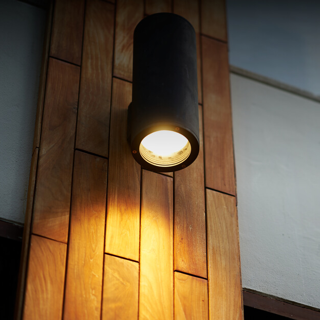 Wall-mounted, cylindrical black lamp reminiscent of landscape lighting in New Orleans, emits a warm glow onto wooden paneling, creating an inviting ambiance.